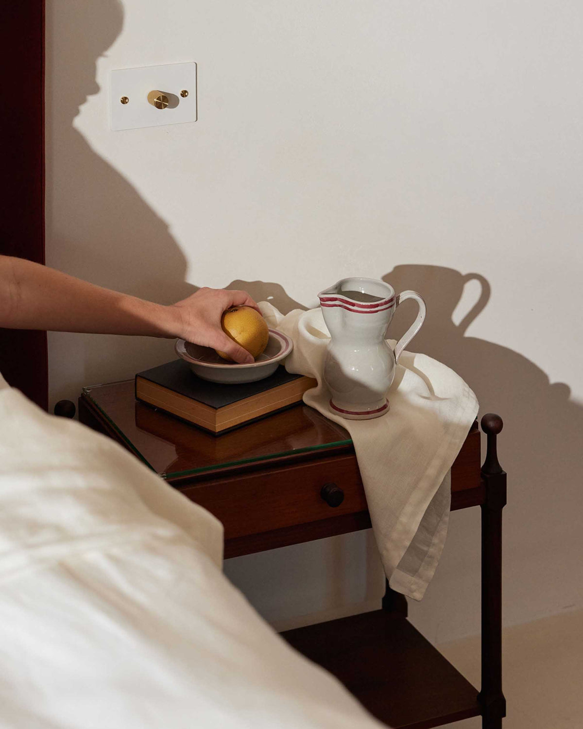A hand reaches for a lemon in a bowl on a bedside table adorned with stacked books, a white pitcher, and Linen Table Napkins - Ivory, neatly folded beside the bed with crisp white bedding.
