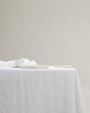 A Linen Tablecloth - White covers a table set with a white cup, a white speckled plate, and a wooden fork, all neatly arranged against a neutral background.