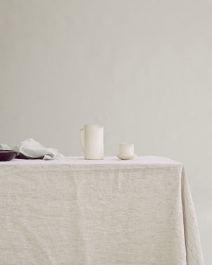 A minimalist scene featuring a ceramic pitcher and cup arranged on the Pinstripe Linen Tablecloth, with a folded cloth and bowl to the left, all set against a neutral background.