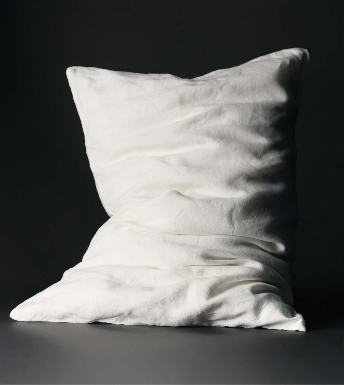 A white, slightly wrinkled pillow sits upright against a dark, plain background. The pillow appears soft and plush, with subtle shadows emphasizing its creases and folds.