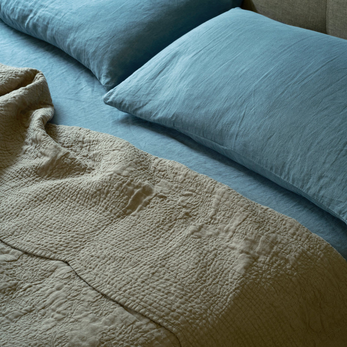 Two light blue pillows rest on a neatly made bed with a blue sheet and a textured beige quilt partially covering it. The image conveys a calm, cozy, and inviting bedroom atmosphere.