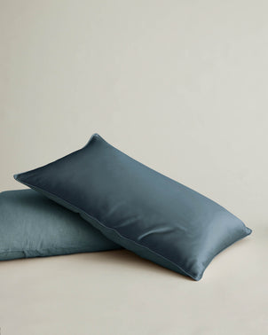 Two blue rectangular pillows, one with the Silk Linen Flip Pillowcase - Bluestone in shiny mulberry silk resting diagonally over a matte pillow, set against a plain light background.