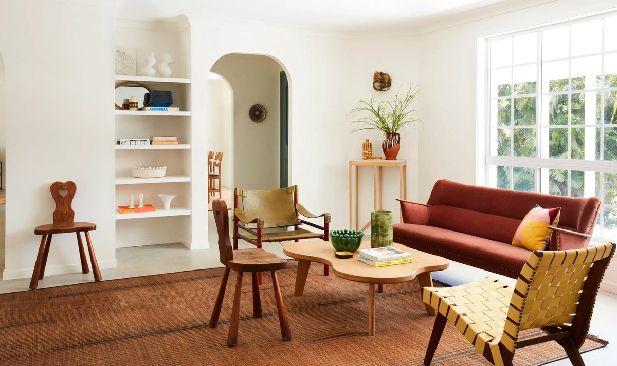 Bright living room with a red sofa, woven chairs, wooden tables, brown rug, bookshelf, potted plant, large window, and arched doorway leading to a dining area. The decor is eclectic and features warm, earthy tones.