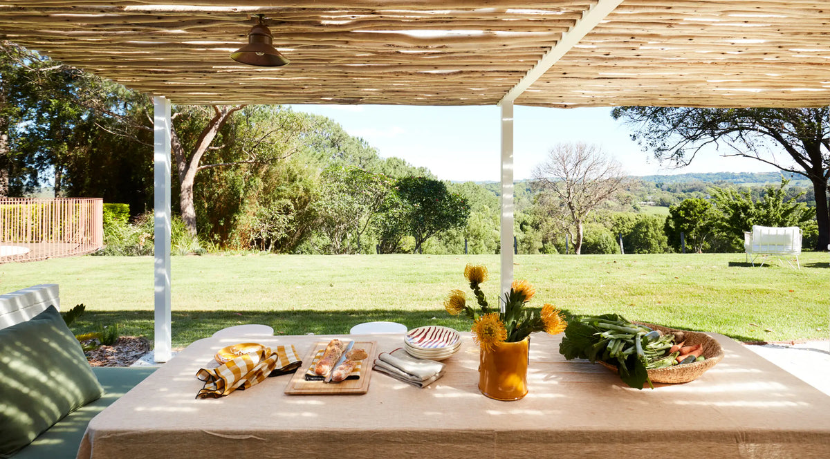 Outdoor dining table set with food, flowers, and vegetables under a wooden pergola, overlooking a green lawn and trees with a scenic landscape in the background.