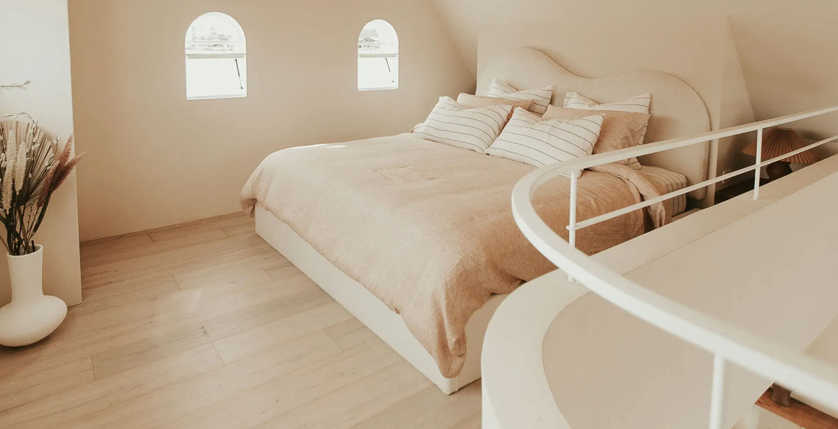 Minimalist beige bedroom with a double bed, striped pillows, light wood floors, three arched windows, and a modern white railing in the foreground. Dried plants in a vase sit in the corner.