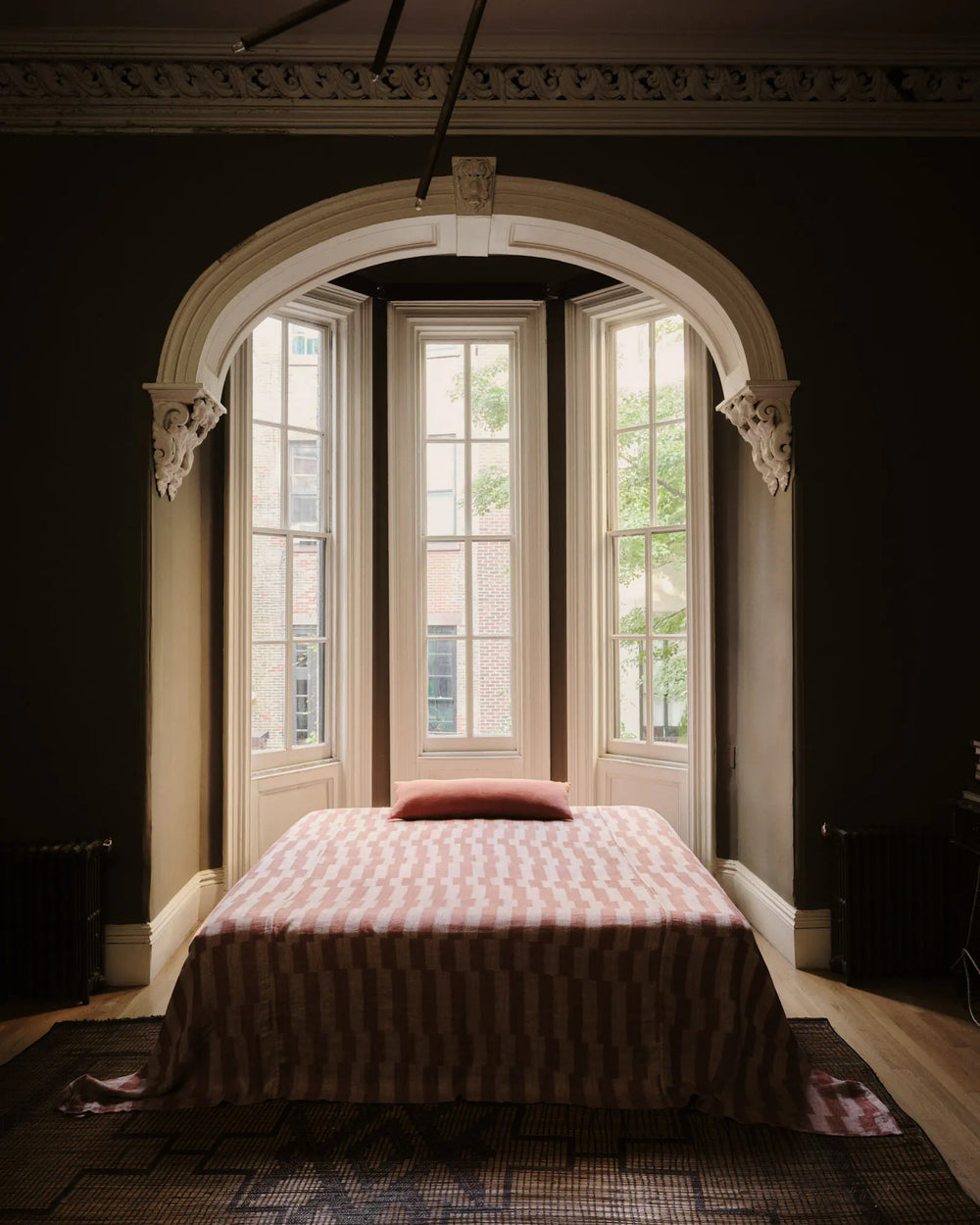 A neatly made bed with a pink patterned blanket and pillow stands before tall bay windows framed by ornate white trim in a dimly lit, elegant room.
