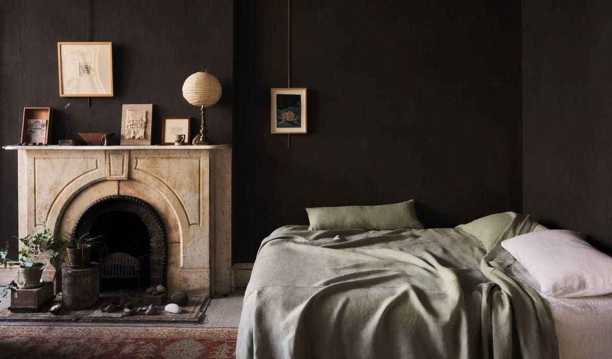 A cozy bedroom with dark walls, a marble fireplace decorated with art and a lamp, and an unmade bed with green bedding and a white pillow. Plants and a small rug add to the warm, lived-in atmosphere.