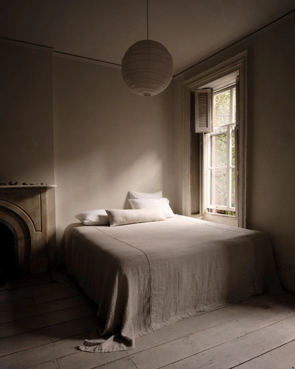 Minimalist bedroom with a bed covered in beige linens, two pillows, soft natural light from a window, light wood floors, a round hanging lamp, and a decorative fireplace mantel on the left.