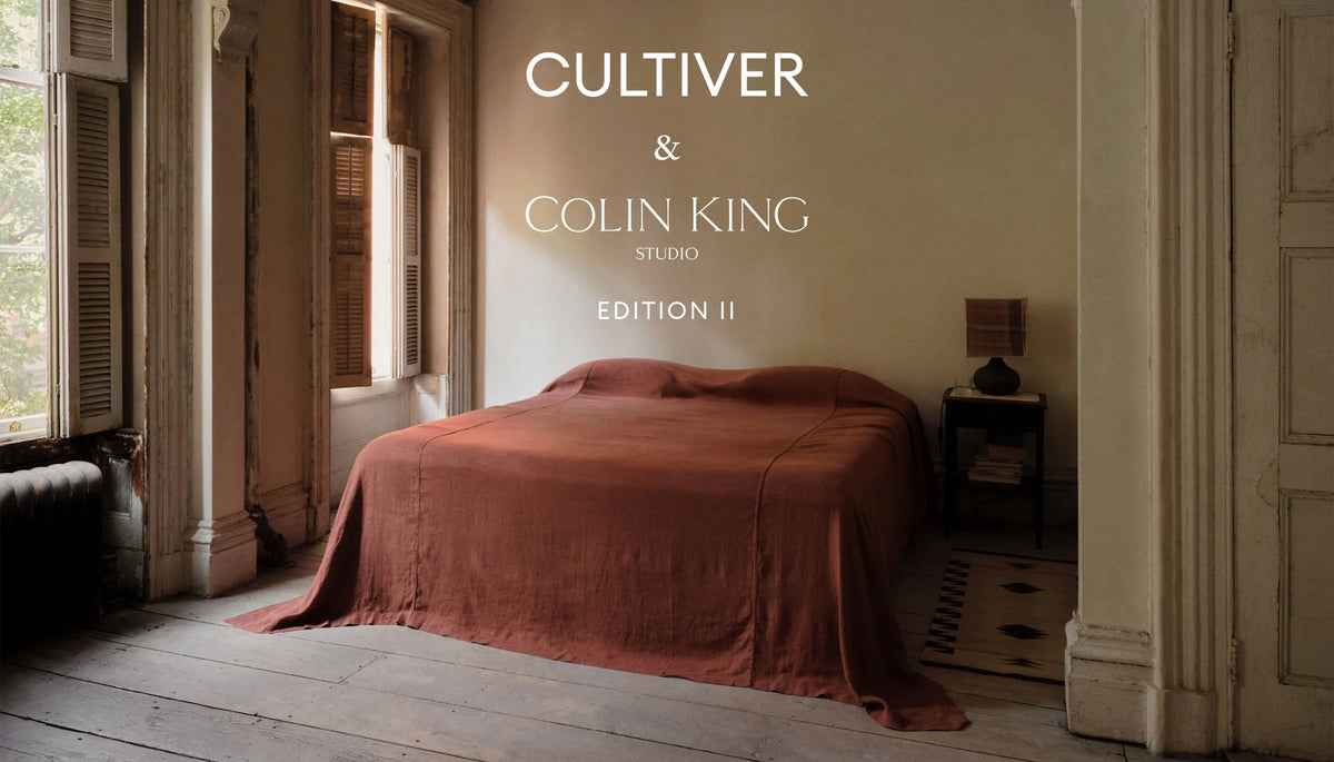 A cozy bedroom with sunlight through tall windows, a bed covered in a rust-colored linen, a side table with a lamp, and text reading CULTIVER & COLIN KING STUDIO EDITION II.