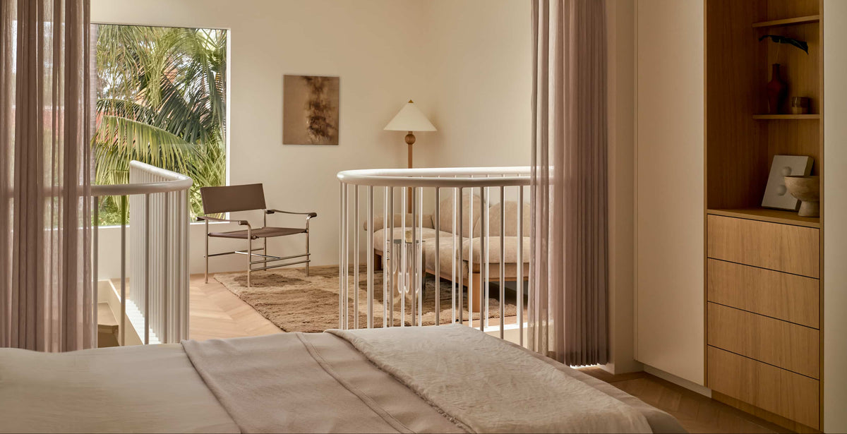 A bright, modern bedroom opens to a cozy sitting area with a chair, sofa, and lamp. Large window shows green palm trees outside. Neutral tones and soft light create a calm atmosphere.