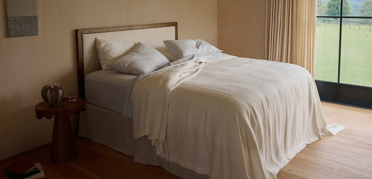 A neatly made bed with cream-colored sheets and pillows sits near a large window with beige curtains, allowing natural light into the room. A small wooden side table holds a glass and decorative items.