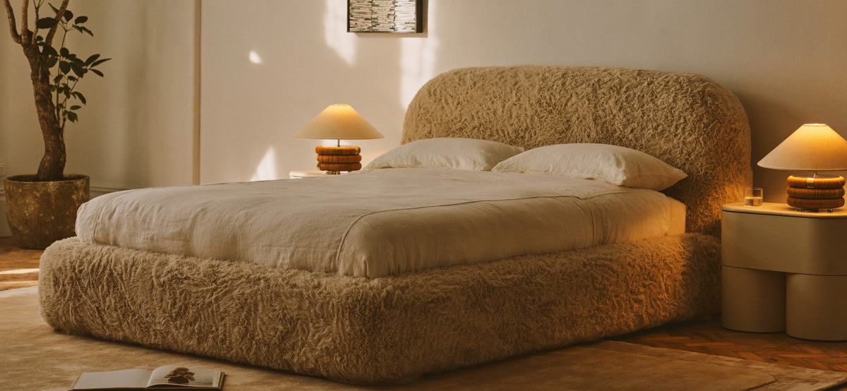 Cozy bedroom with a fluffy beige bed, white bedding, two bedside tables with lamps, a potted plant, and warm sunlight streaming through the window.