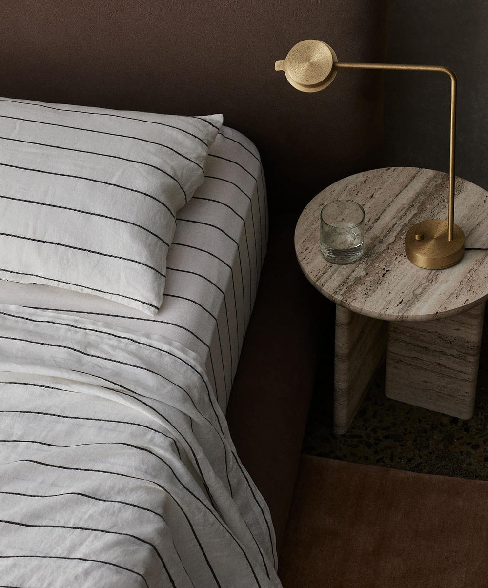 A neatly made bed with white and black striped sheets is next to a small marble nightstand holding a glass of water and a gold modern lamp.