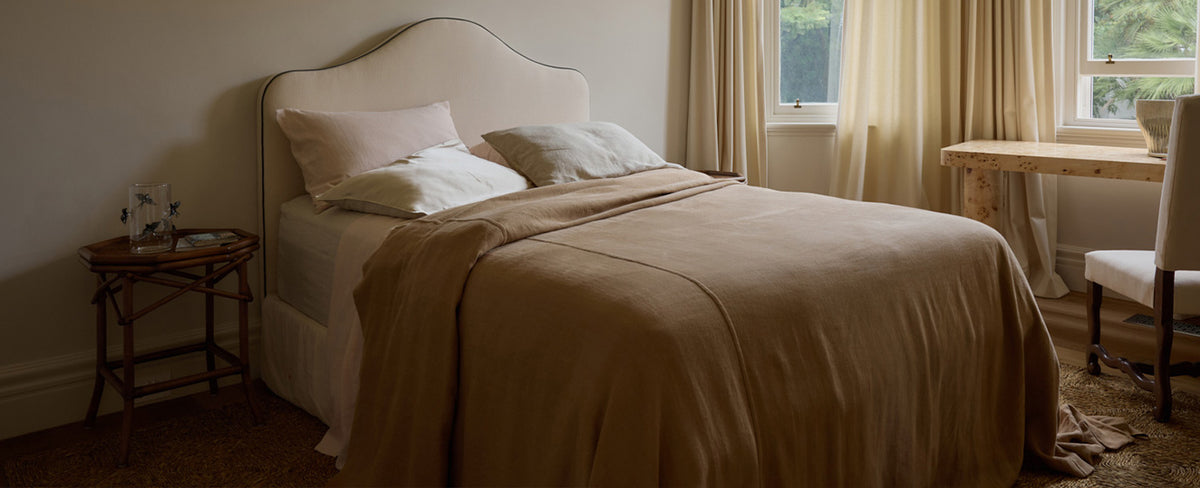 A neatly made bed with beige bedding sits near a window with light curtains. A small wooden table and chair are nearby, and sunlight filters into the cozy, softly lit bedroom.