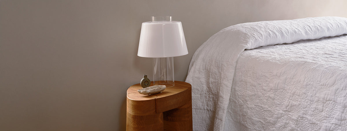 A wooden bedside table with a modern white lamp, a small dish, and a perfume bottle next to a neatly made bed with a white quilted cover. The background wall is plain and light-colored.