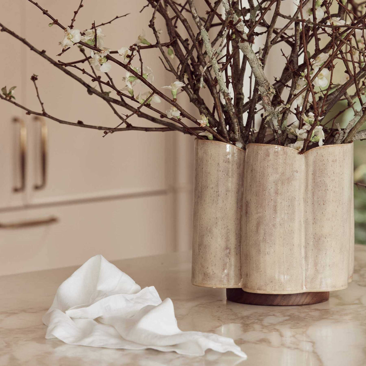 A beige ceramic vase with budding branches sits on a marble countertop beside a crumpled white cloth, with soft natural light in the background.