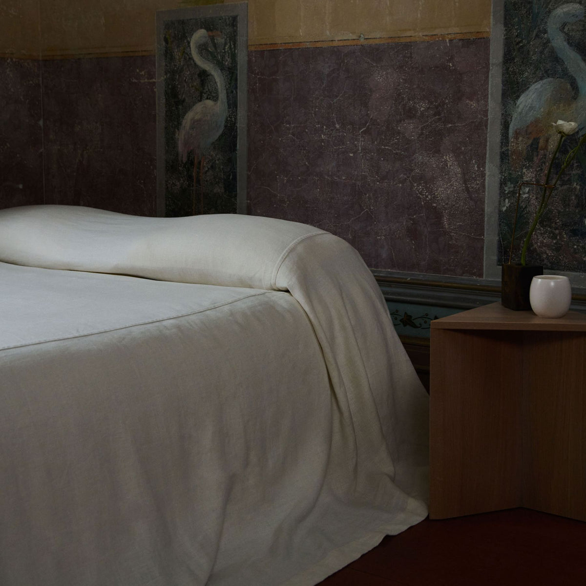 A neatly made bed with a white sheet and blanket is next to a small wooden nightstand holding a white vase with a single flower. The wall behind features artwork of white birds.