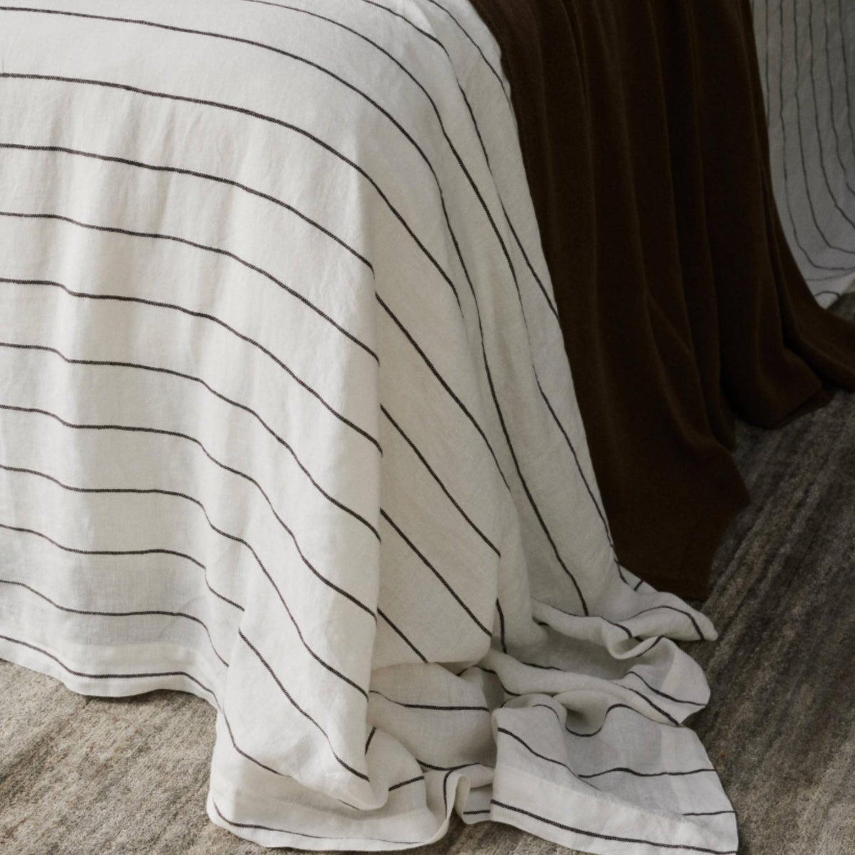 A bed with a white blanket featuring thin black stripes draped over it, next to a solid dark brown blanket, both touching a textured beige carpeted floor.