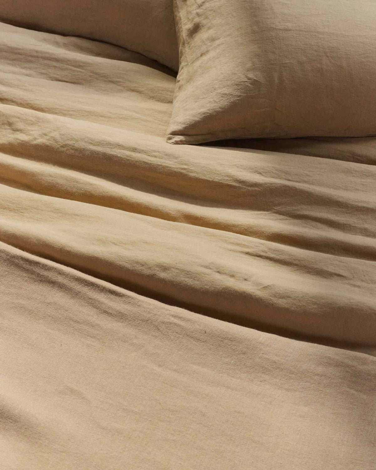 Beige linen bedding with a pillow and slightly wrinkled sheets. The fabric has a soft, textured appearance and a neutral, calming color. The scene gives a cozy, inviting feel.