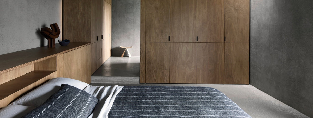 Minimalist bedroom with gray walls, wooden built-in cabinets, a bed with striped bedding, and a small stool in the background. The room has a calm, modern, and uncluttered design.