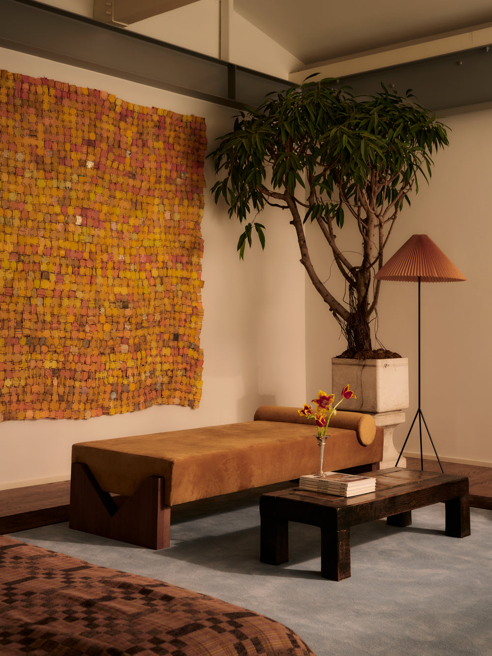 A cozy room with a tan daybed, a dark wood coffee table holding flowers, a large leafy potted plant, a pleated lamp, and a colorful textured wall hanging in yellow and pink tones.