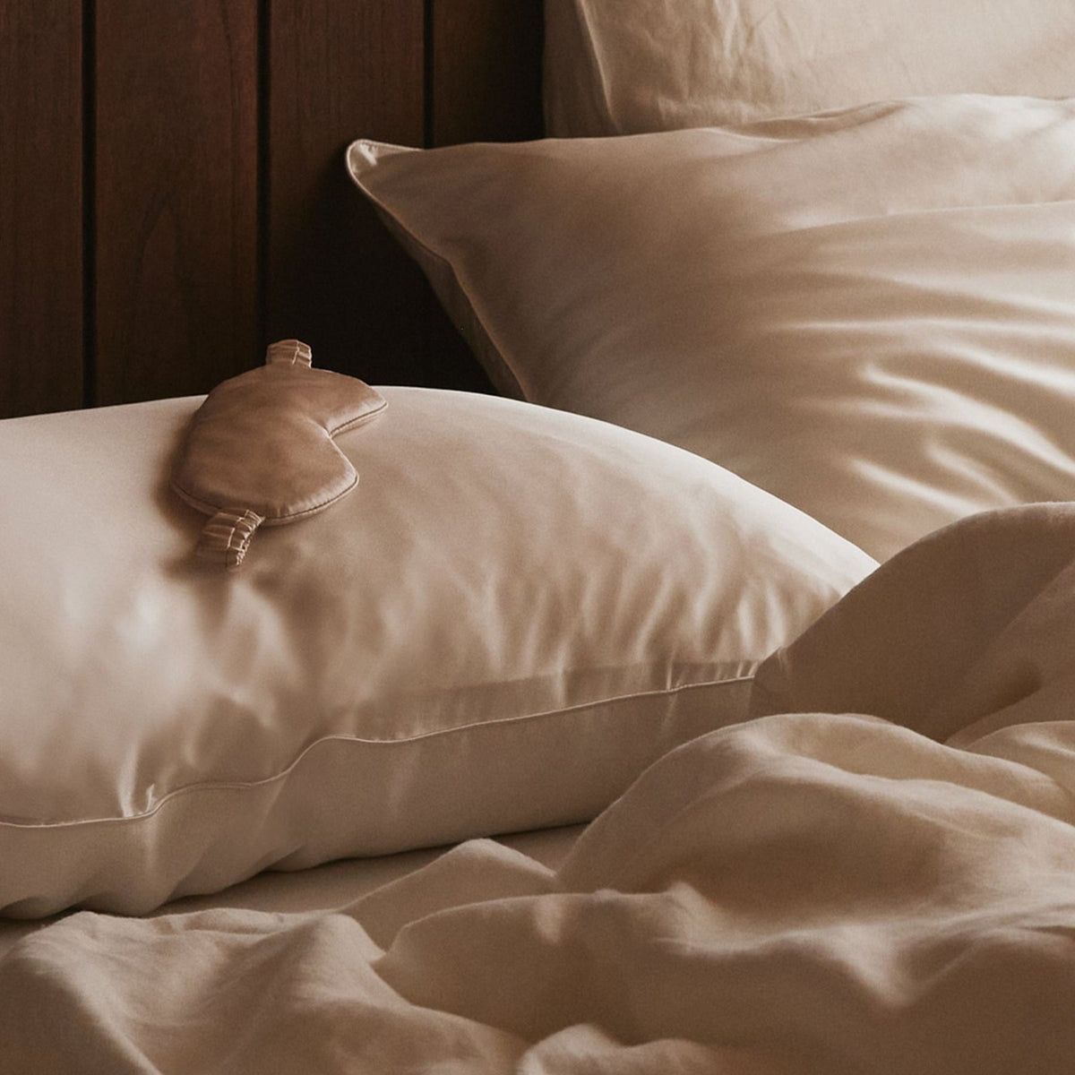 A beige sleep mask rests on a white pillow atop an unmade bed with creamy, rumpled sheets near a wooden headboard.