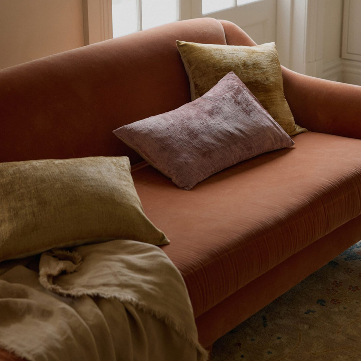 A rust-colored sofa with three textured pillows in sage green and mauve, plus a beige throw blanket, sits near a bright window, creating a cozy and inviting atmosphere.