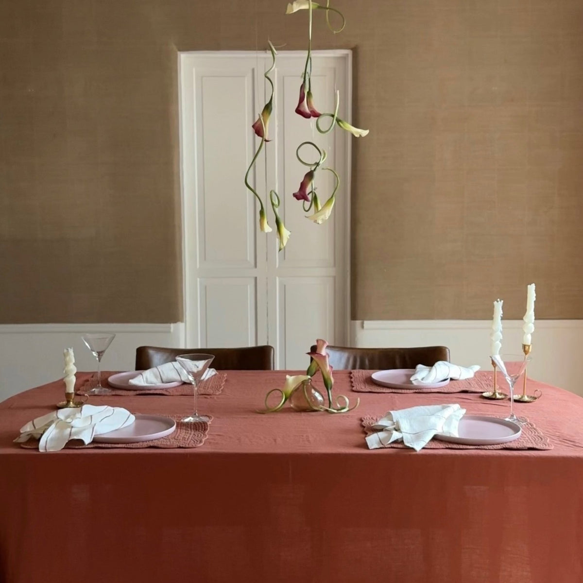 A dining table with a rust-red tablecloth, minimal place settings, twisted napkins, and tall twisted white candles. Curled calla lilies hang above the table against a neutral wall and white double doors.