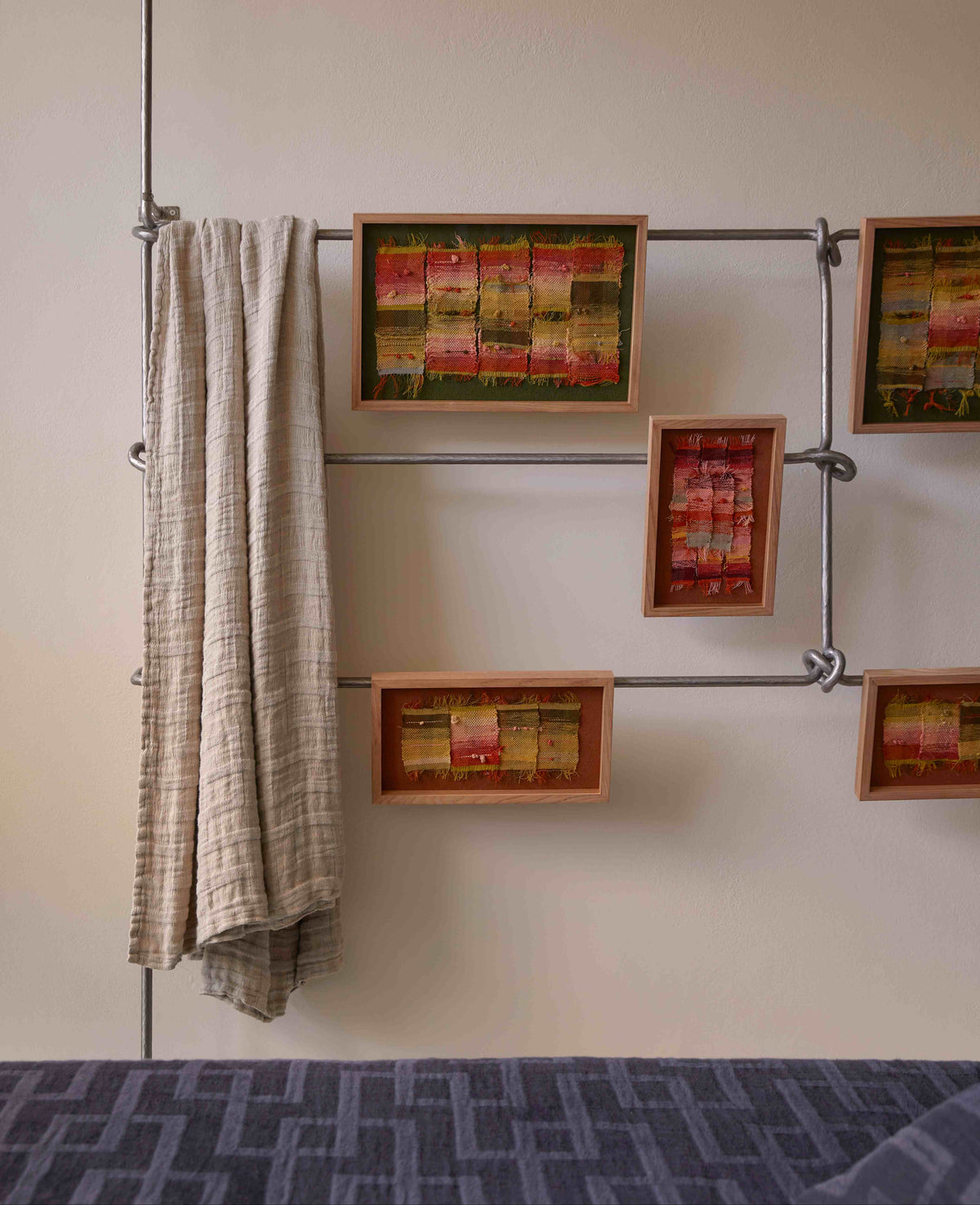 A beige textured blanket hangs on a metal pipe rack mounted to a wall, alongside five framed abstract paintings in red, yellow, and green tones. A blue patterned bedspread is visible in the bottom foreground.