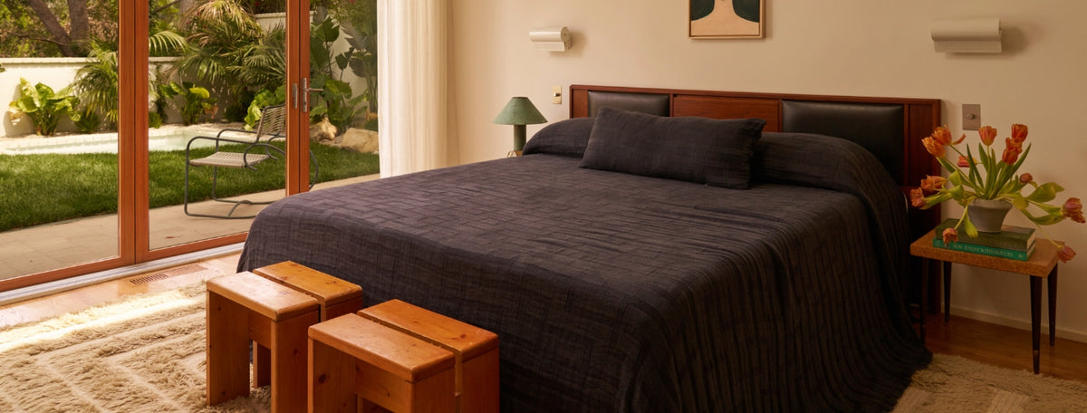 A modern bedroom with a large bed covered in a dark quilt, wooden stools at the foot, a side table with tulips, and glass doors opening to a garden with greenery and a lounge chair.