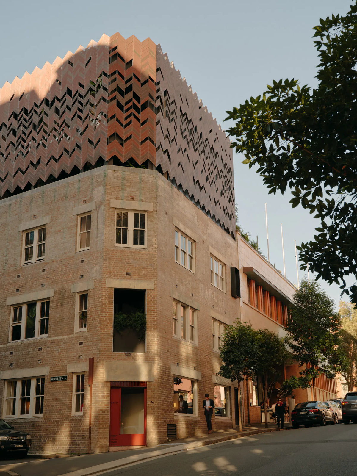 A modern building with a brick lower section and a zigzag-patterned upper facade stands on a street corner. People walk nearby, and trees line the street under a clear sky.
