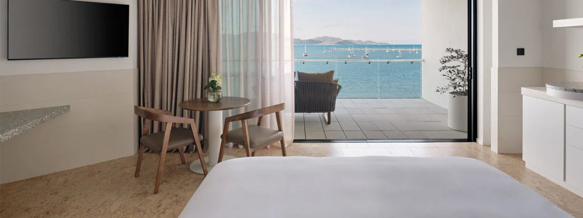 A modern hotel room with a large bed in the foreground, a small dining table and chairs, a wall-mounted TV, and floor-to-ceiling glass doors opening to a balcony overlooking a blue sea.