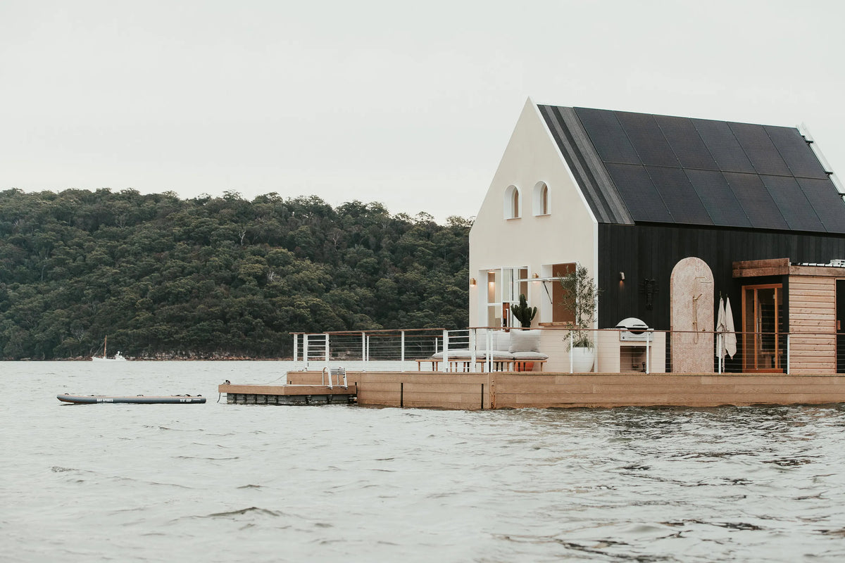 A modern house with large windows and a deck sits on the water, surrounded by calm waves. Trees cover the distant shoreline under an overcast sky.