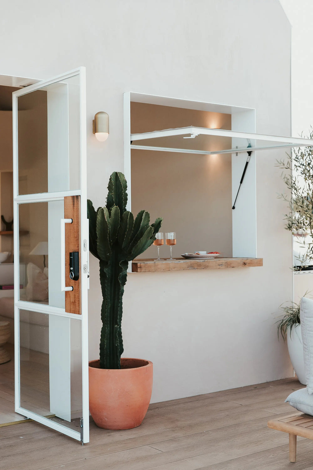 A tall cactus in a terracotta pot stands next to an open glass door and a serving window with drinks on a wooden counter, in a modern, minimal patio setting.