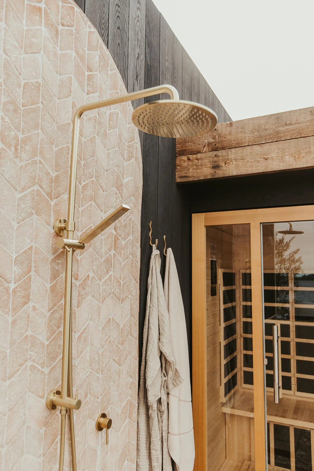 Outdoor shower with a round brass showerhead on a tiled wall, two striped towels hanging on hooks, and a wooden sauna with glass door visible in the background.
