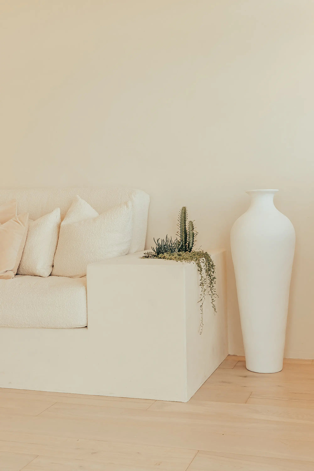 Minimalist living room with a cream-colored sofa, beige pillows, a built-in planter with green succulents, and a large white vase on a light wooden floor against a soft beige wall.
