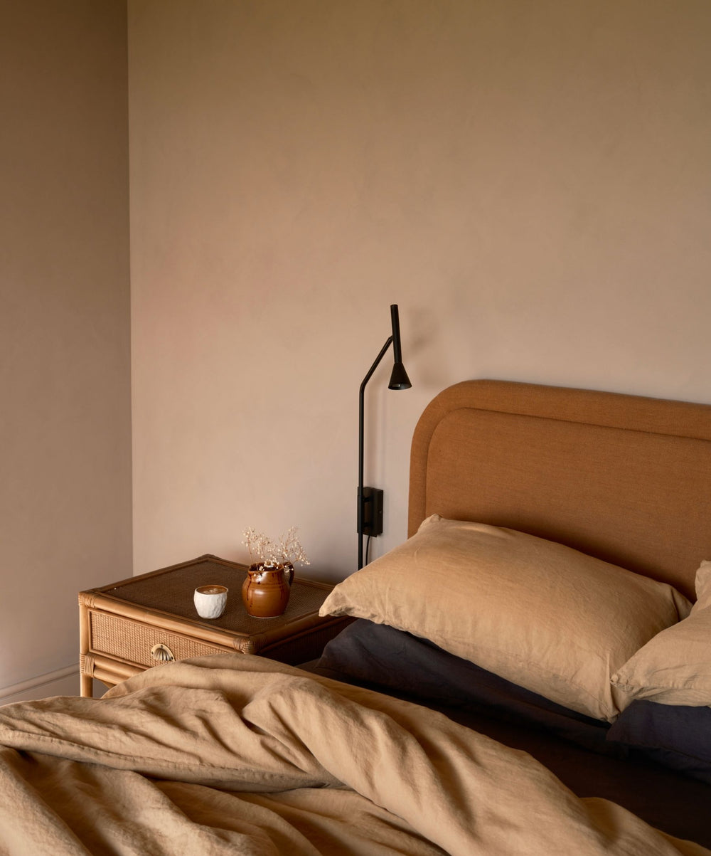 A cozy bedroom with a tan upholstered headboard, beige bedding, a wicker nightstand, a black reading lamp, a brown vase with dried flowers, and a small white cup, all set against a beige wall.