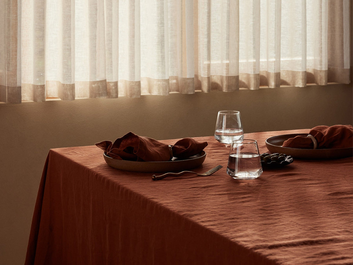 A sunlit table with an orange cloth, two plates with brown napkins, two glasses of water, a fork, and grapes, beside sheer curtains.