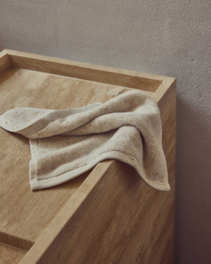 A Woven Speckle Cotton Wash Cloth in beige is draped over the edge of a light brown stone countertop, set against a textured, light-colored wall.