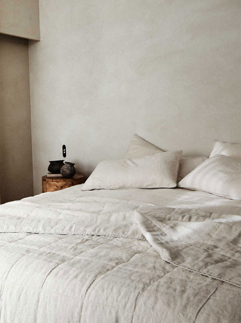 A neatly made bed with beige linens and three pillows, next to a small wooden nightstand holding two ceramic items, against a plain light-colored wall.