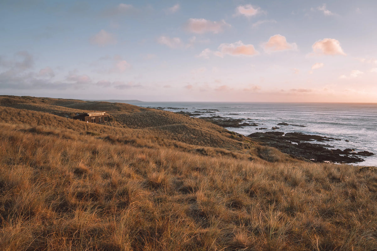 A small cabin sits on a grassy hill overlooking a rocky coastline and calm ocean under a pastel sky at sunset.