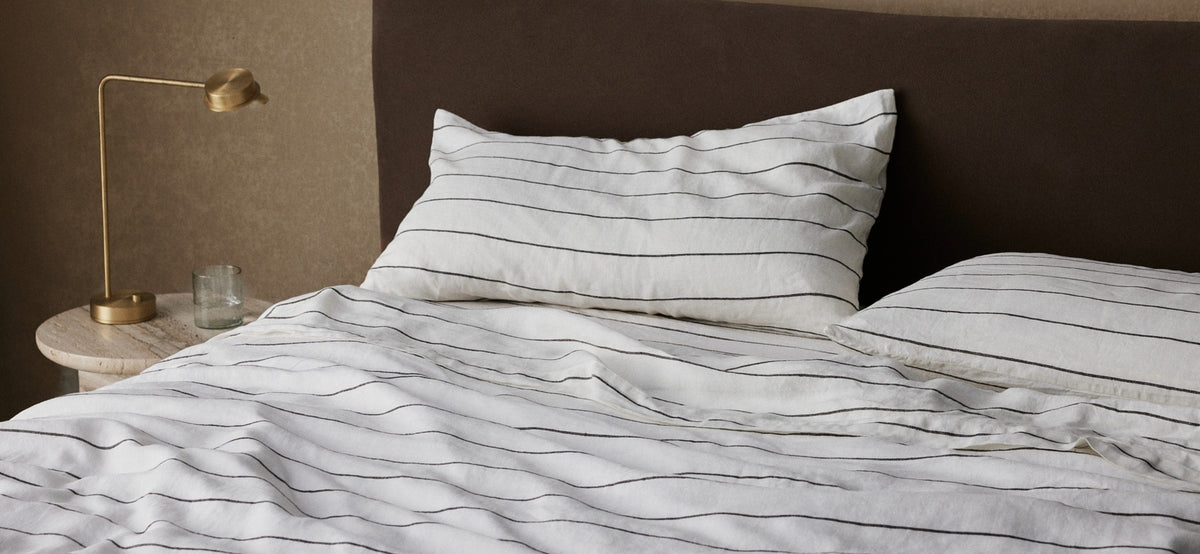 A neatly made bed with white and black striped bedding, two pillows, a small round bedside table with a glass, and a gold lamp against a brown upholstered headboard.
