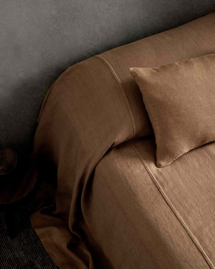 A neatly made bed with brown linen sheets, the Heavyweight Linen Bedcover in Sand, and a matching pillow against a gray wall, viewed from above.