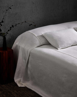A neatly made bed with a white Heavyweight Linen Bedcover and a single pillow sits beside a dark wooden nightstand holding a black vase with a minimalist branch, set against a textured gray wall.