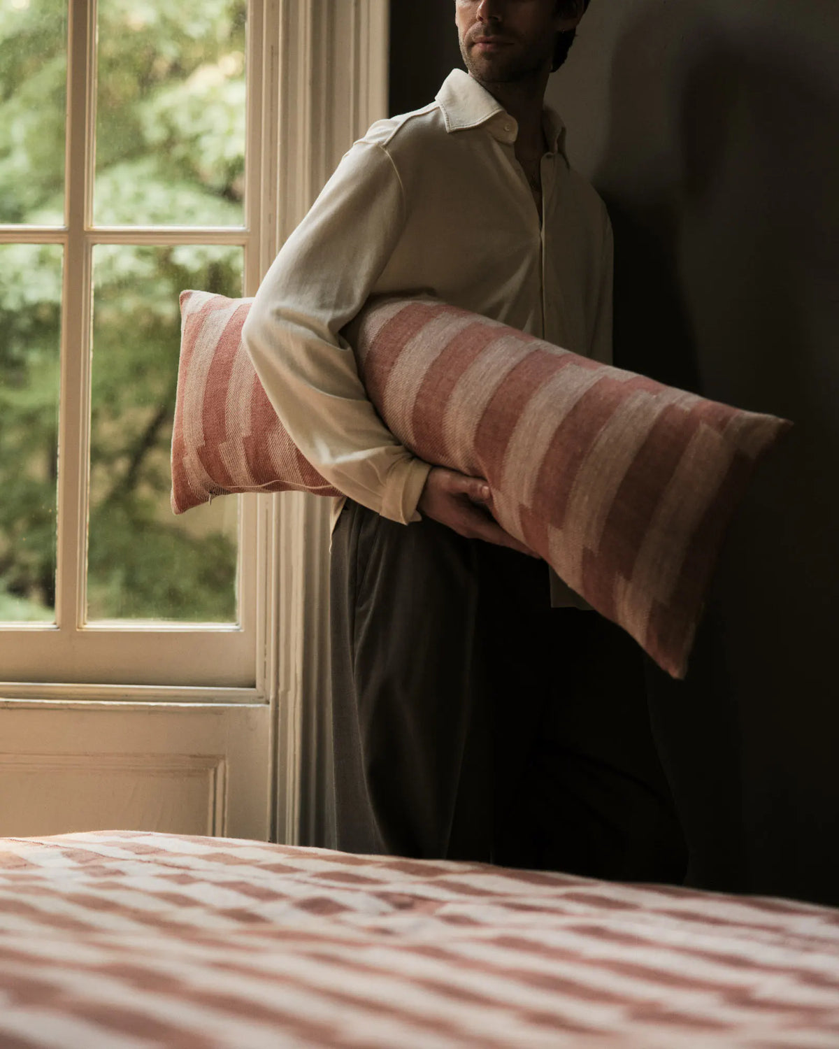 A person in a cream shirt holds a long, striped pink pillow. They stand by a sunlit window, with trees visible outside. A matching pillow is on a nearby bed.