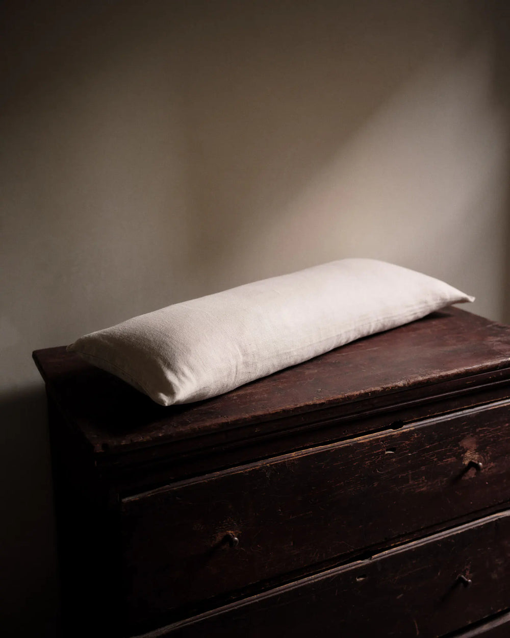 A rectangular white pillow rests on top of a dark, worn wooden dresser in a softly lit room with neutral walls.