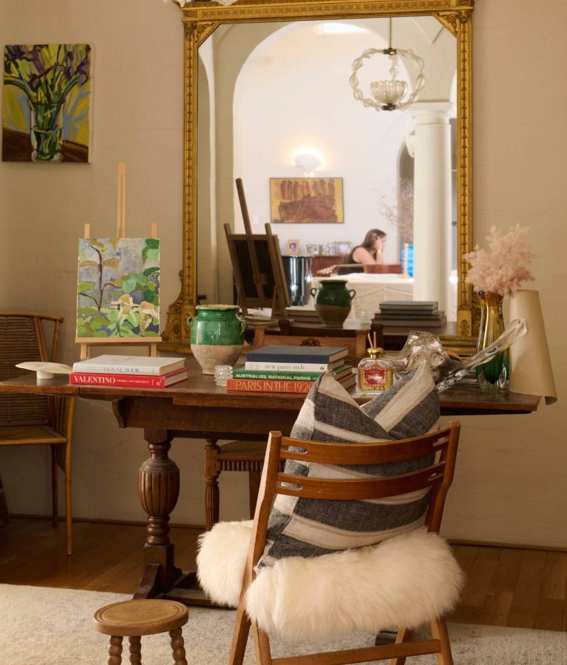A cozy art-filled room with a wooden table, books, green vases, and a painting. A striped pillow and fluffy white cushion are on a chair. A large gold mirror reflects a woman sitting at another table.