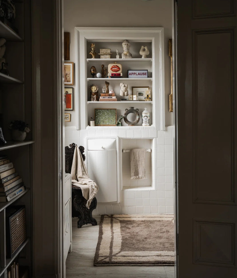 A cozy bathroom with white tiles, a brown rug, a towel rack, ornate chair, and shelves filled with books, small statues, framed art, and decor such as an APPLAUSE sign and gold figurines.