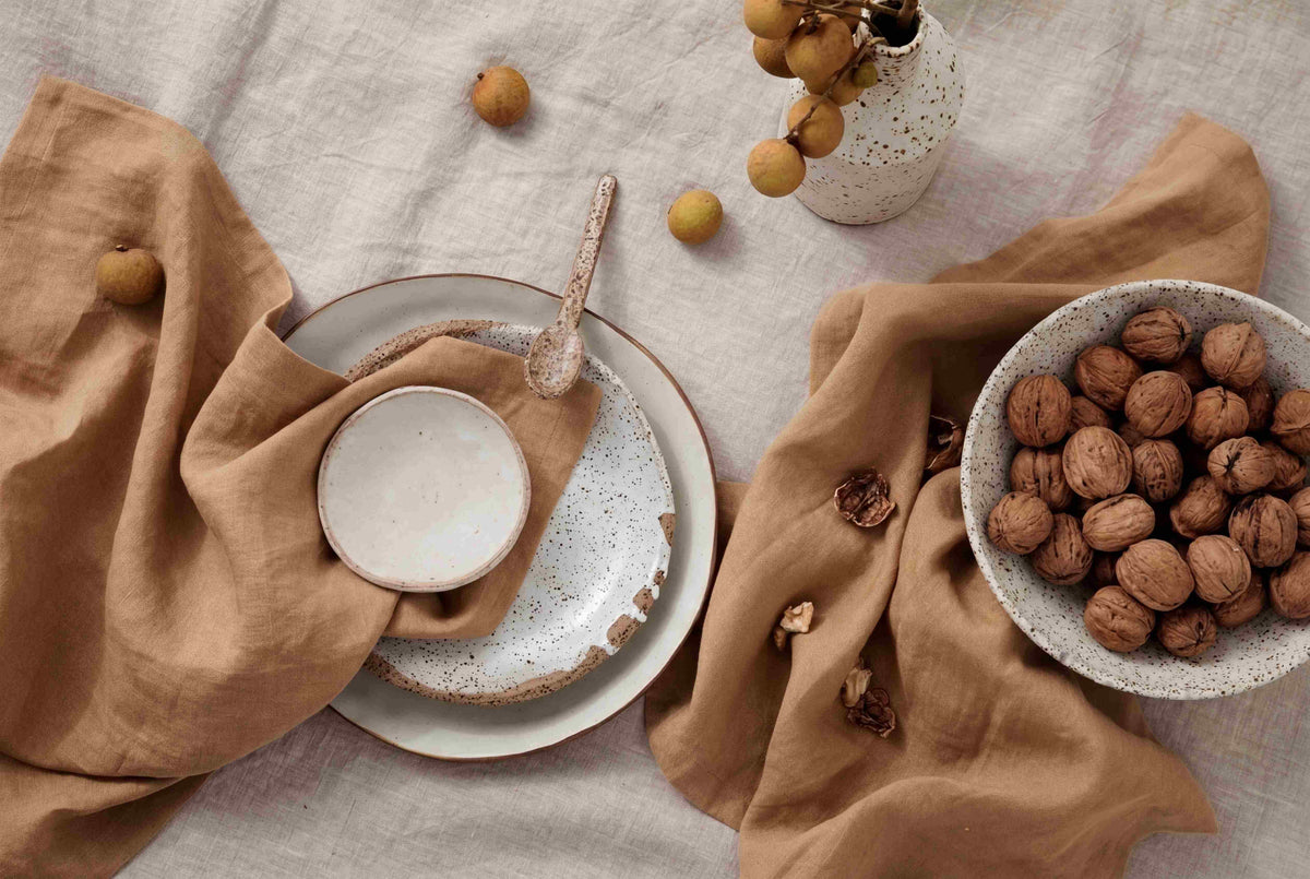 Table setting with neutral-toned ceramic plates, a bowl of walnuts, brown linen napkins, a rustic spoon, and a small vase with dried branches, all on a beige tablecloth.