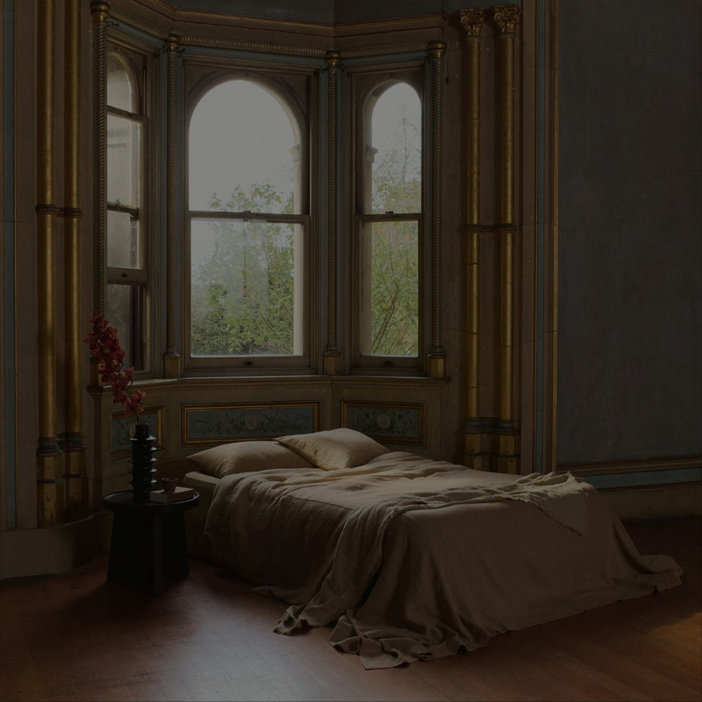 A cozy bed with beige bedding sits near tall, arched windows in a room with ornate gold trim and wooden floors. A small round table holds a vase with pink flowers beside the bed.